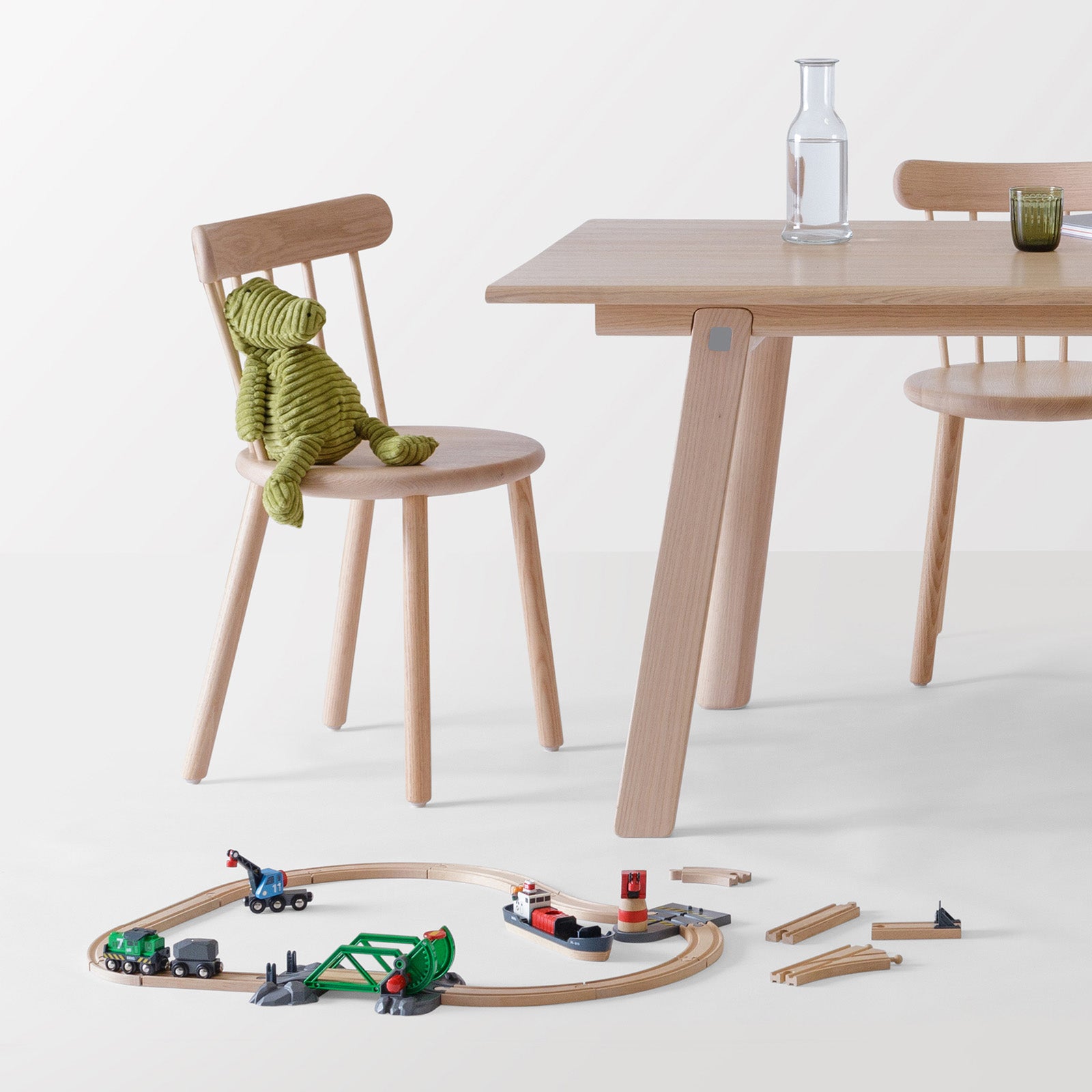 Ai Chair in natural oak paired with the T&B Table, styled with a green knitted teddy alligator and a wooden toy train set in a playful and minimalist dining setup.