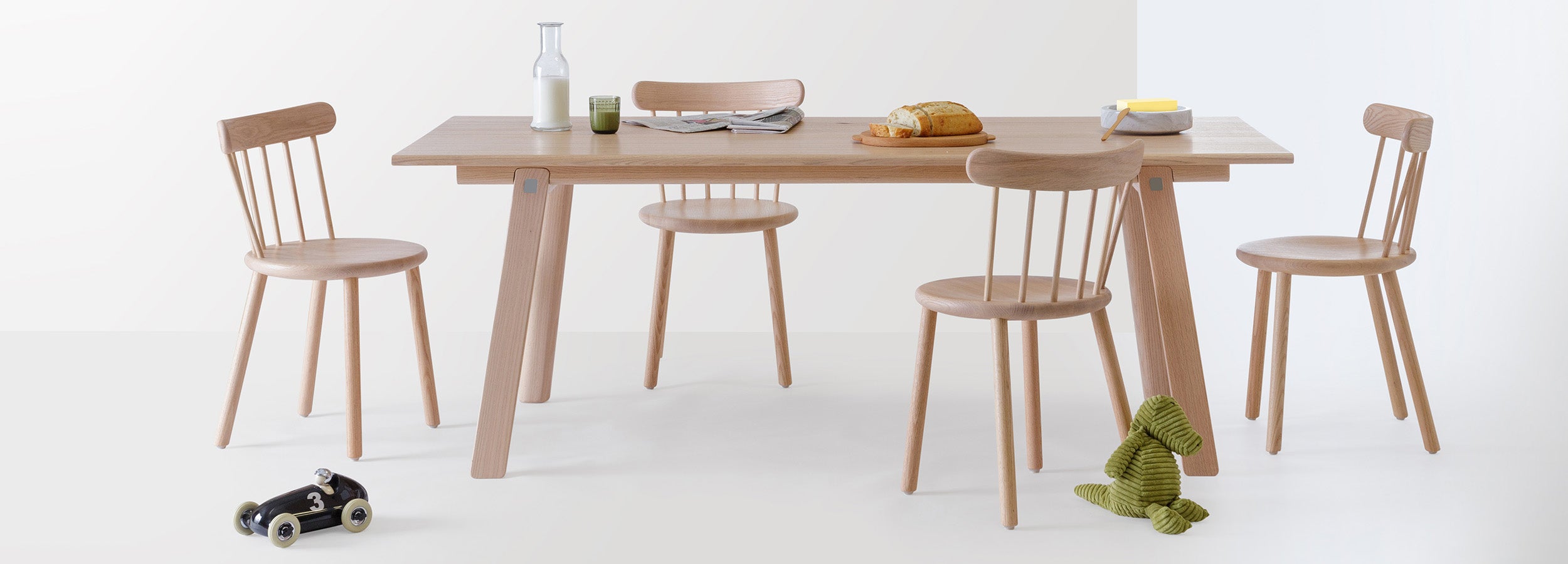 Ai Chairs in natural oak paired with the T&B Table, styled for a relaxed breakfast setting with bread, milk, and a newspaper in a bright, minimalist dining room.
