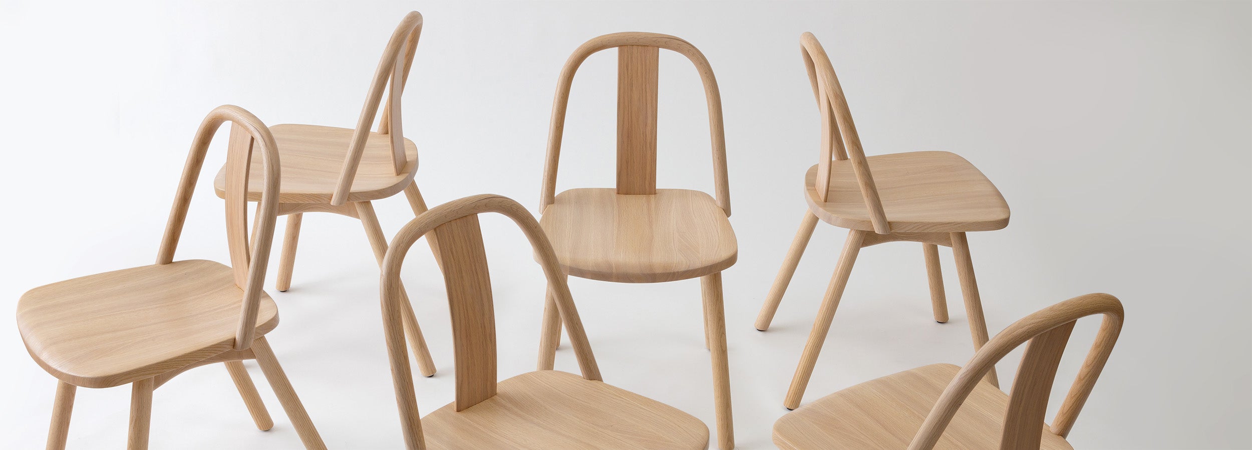 A collection of Chin-Chin chairs crafted in natural oak, showcasing their minimalist curved design and versatile aesthetic for dining or gathering spaces.