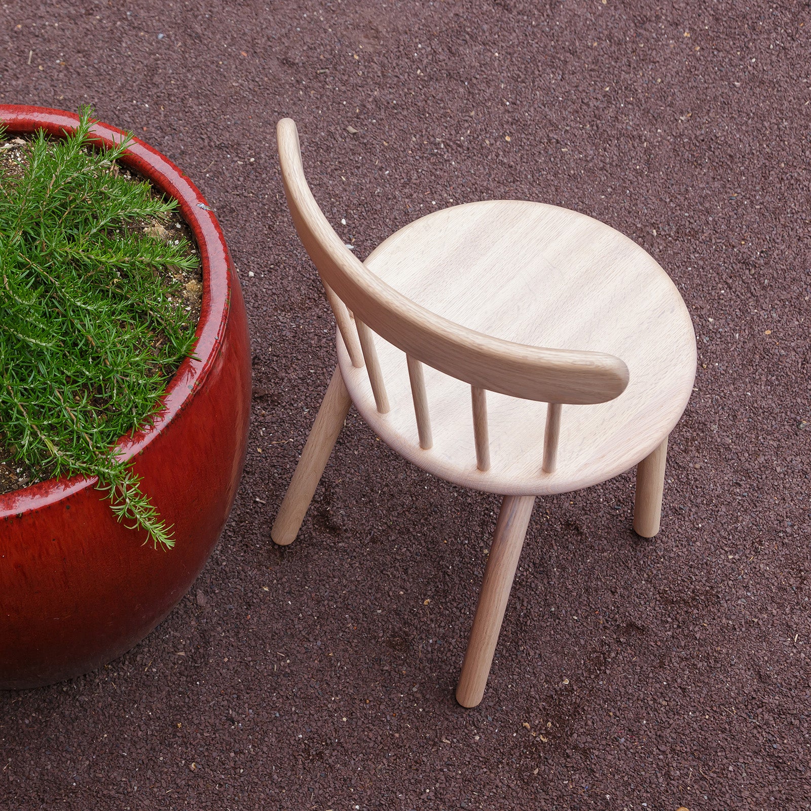 Ai Chair in natural oak placed outdoors on a gravel surface, next to a large red planter filled with greenery, showcasing its versatile design.