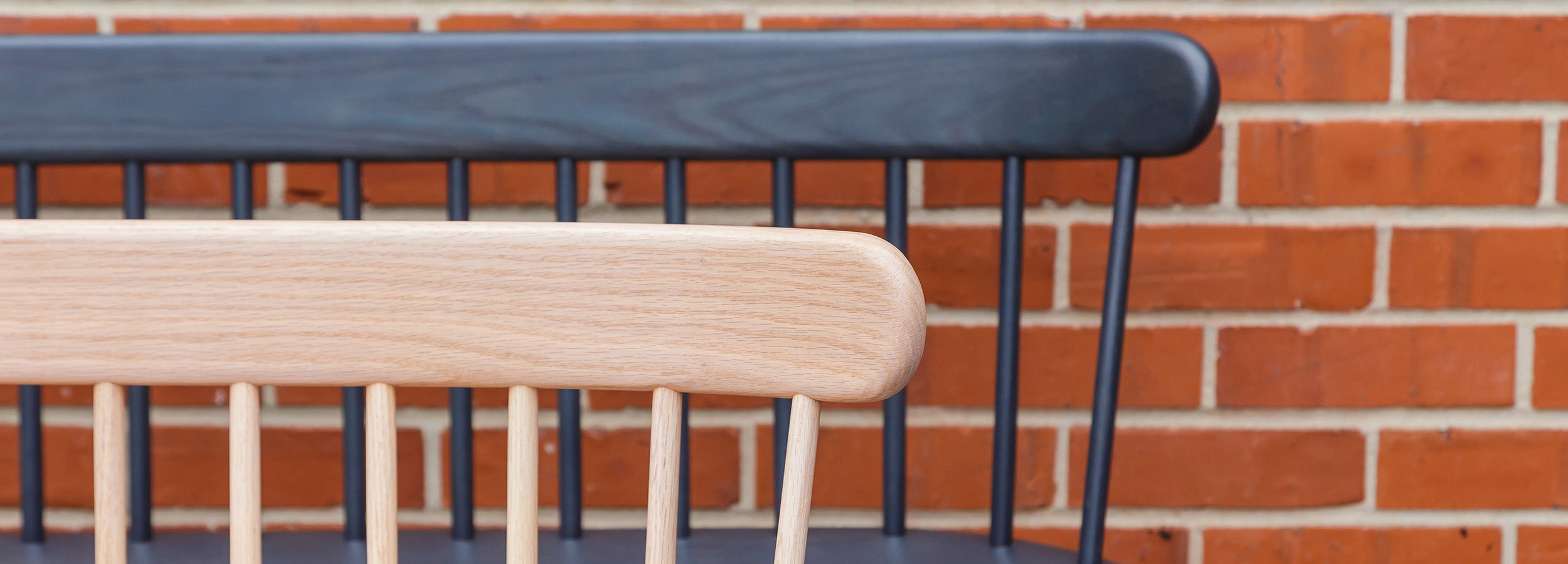 Close-up of Ai Settee in natural oak and Ai Chair in black-blue finish, showcasing the spindled backrests and contrasting tones against a red brick wall.