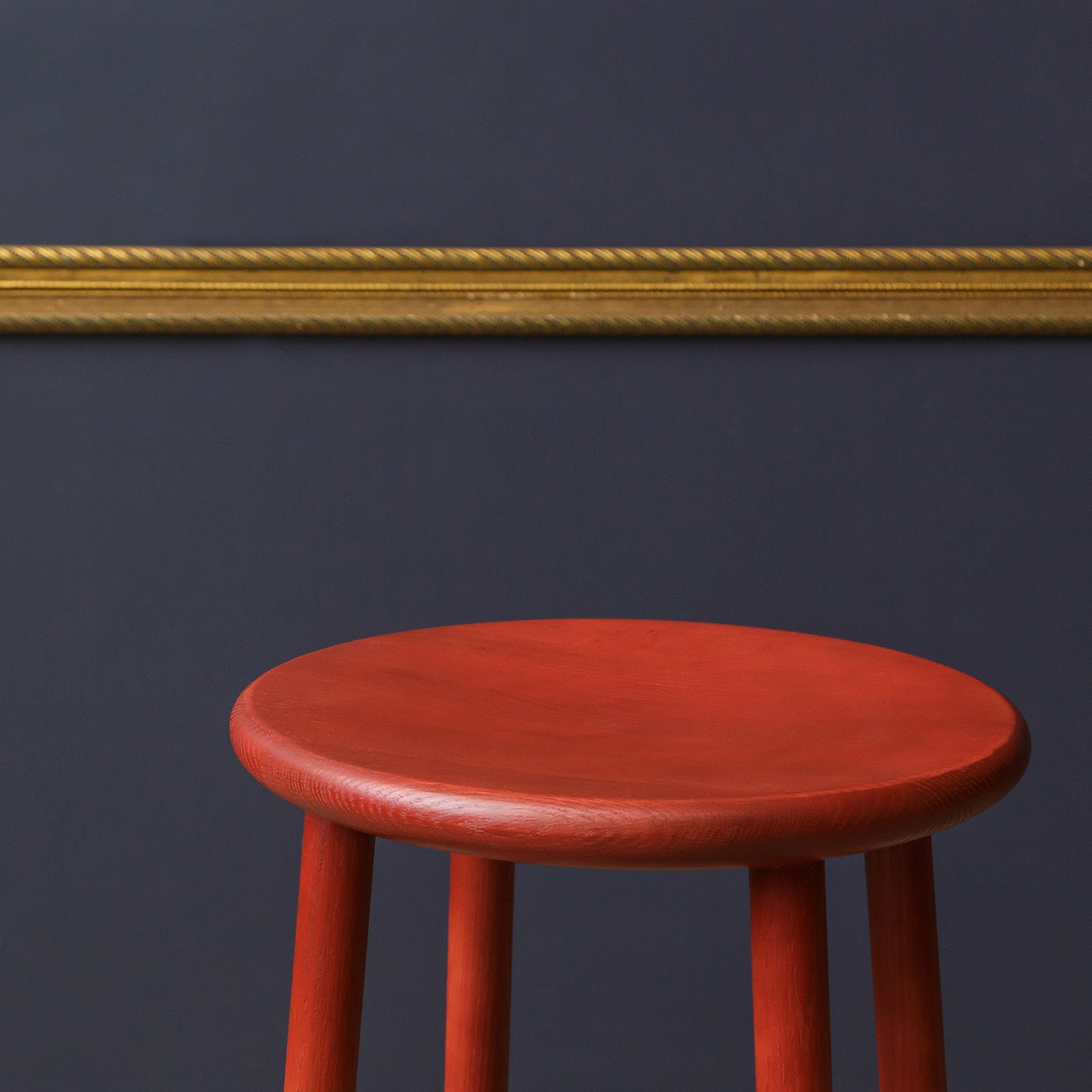 Close-up of the Ai Stool in deep red finish, highlighting its smooth round seat and precise craftsmanship against a navy wall with a gold trim.