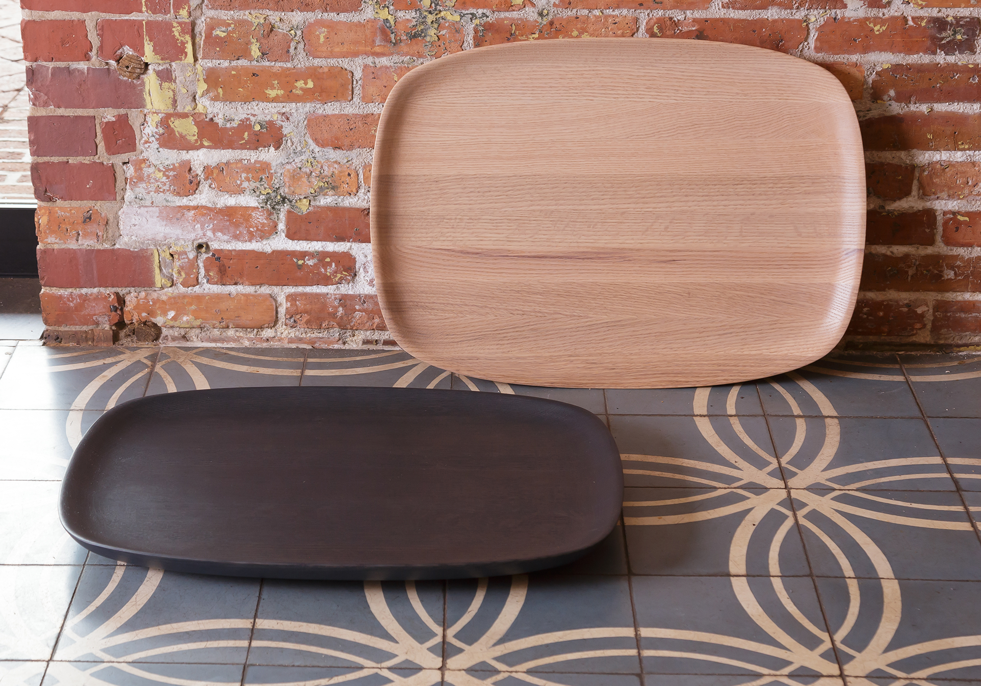 Kokoro Trays in natural oak and black-blue displayed on a tile floor and leaning against a wall, showcasing their versatile design and fine craftsmanship. Suitable for use on tables or as floor décor.